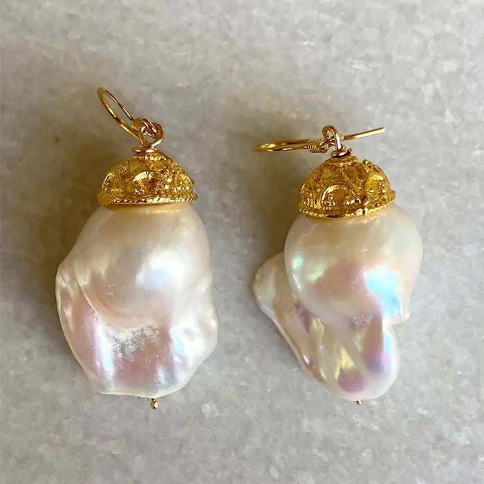 BITTERSWEET DESIGNS - CAPPED IVORY BAROQUE PEARL EARRINGS: Elegant, iridescent ivory baroque pearls with ornate gold caps and hoops from BITTERSWEET DESIGNS are displayed on a light gray textured surface.