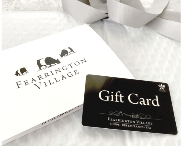 A FEARRINGTON VILLAGE GIFT CARD sits on a white envelope with the brand’s three-cow logo and a soft gray ribbon in the background. Use it at FEARRINGTON VILLAGE favorites like the Spa or Fearrington House Inn.