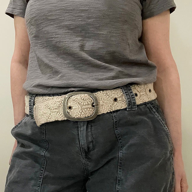 The JENNY KRAUSS - DESSERT DREAMS BELT by JENNY KRAUSS is a beige, hand-embroidered Peruvian belt with intricate floral motifs, metal eyelets, and a silver buckle, artfully coiled on a light surface.