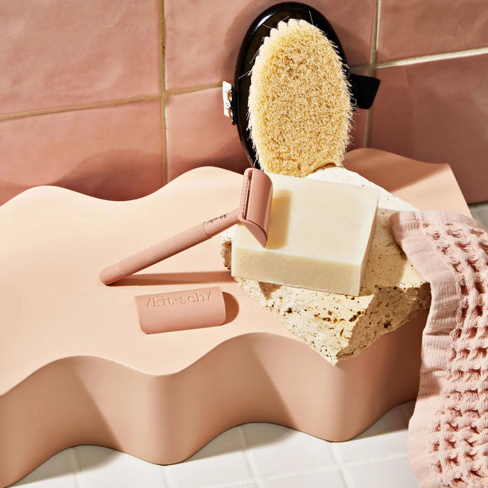 A KITSCH - Shave Butter Bar rests on a marble block, promising the smoothest shave. The packaging emphasizes "All Natural," "pH Balanced," and "Vegan." Below are rosemary, argan nuts, and shea butter, highlighting its plastic-free shaving commitment.