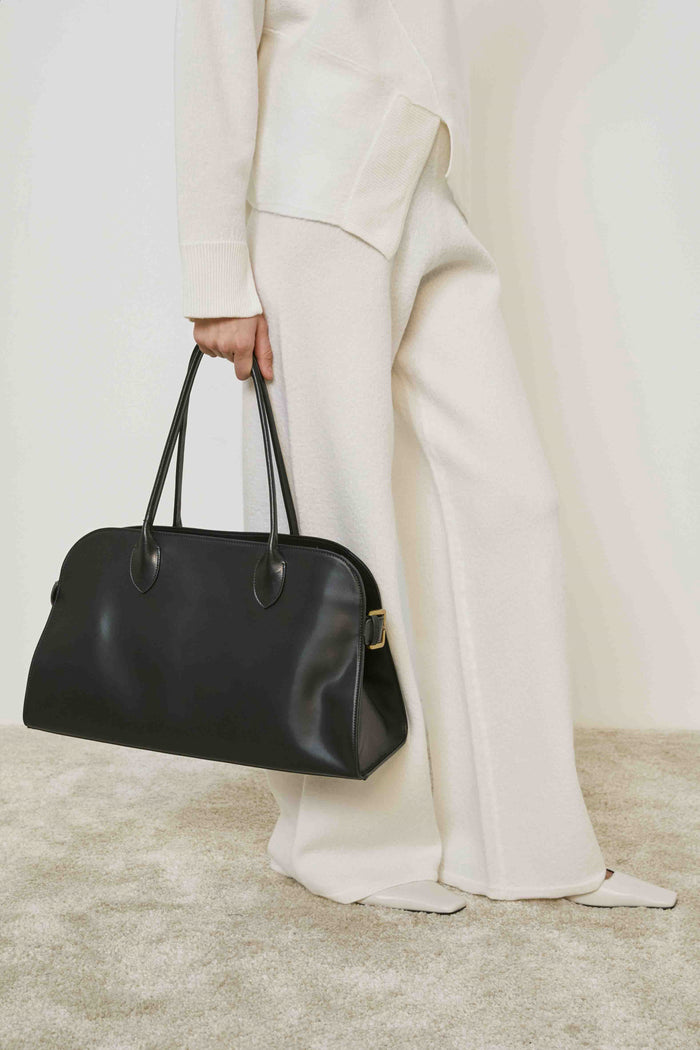 A person in loose cream pants and a matching sweater stands on carpet, holding the MEIMEIJ - BLACK LEATHER HANDBAG by its handles, adding a structured silhouette to the relaxed MEIMEIJ ensemble.