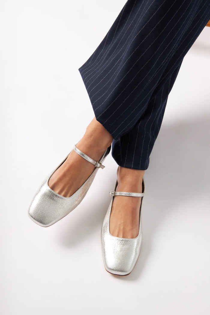 The AUGUSTA - JULIETA MARY JANE by AUGUSTA is a black leather Mary Jane flat with a thin strap and buckle, light brown sole, and subtle square toe, handcrafted in Spain and shown on a light gray background.