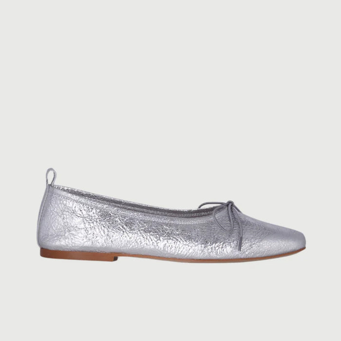 The AUGUSTA - SIENNA BALLET FLAT by AUGUSTA, handcrafted in Spain, showcases a silver finish with a front bow, low brown sole, and heel pull tab, displayed in side profile against a plain light background.