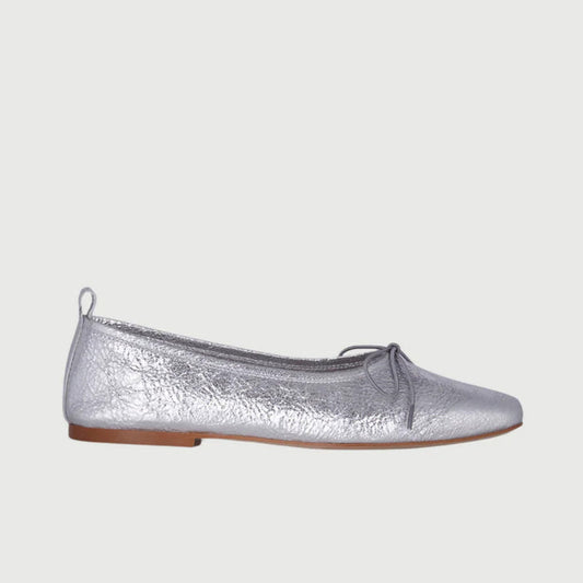 The AUGUSTA - SIENNA BALLET FLAT by AUGUSTA, handcrafted in Spain, showcases a silver finish with a front bow, low brown sole, and heel pull tab, displayed in side profile against a plain light background.