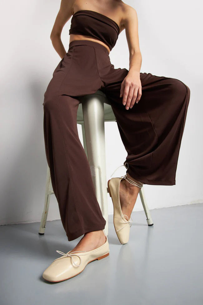 The AUGUSTA - SIENNA BALLET FLAT by AUGUSTA, handcrafted in Spain, showcases a silver finish with a front bow, low brown sole, and heel pull tab, displayed in side profile against a plain light background.