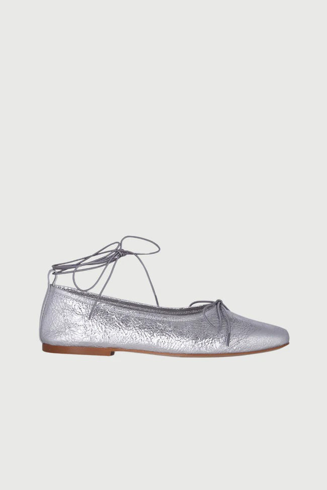 The AUGUSTA - SIENNA BALLET FLAT by AUGUSTA, handcrafted in Spain, showcases a silver finish with a front bow, low brown sole, and heel pull tab, displayed in side profile against a plain light background.