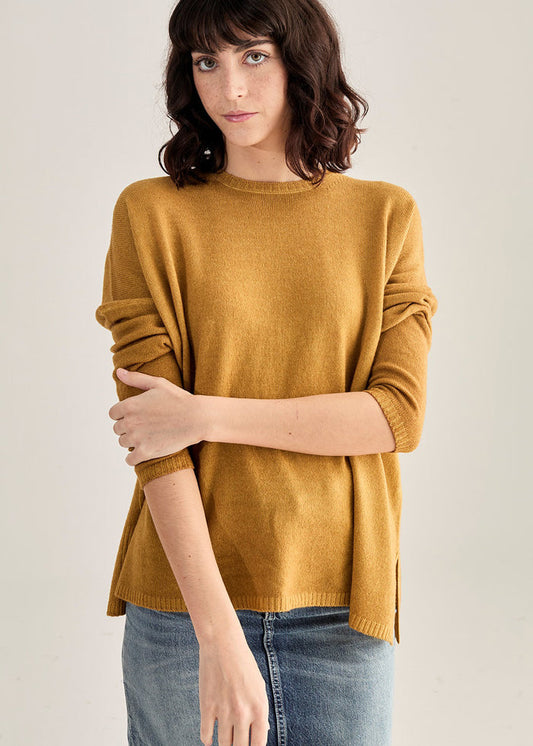 A woman with wavy dark hair wears the BASE MILANO - BOXY OVERSIZED CREWNECK SWEATER in mustard yellow and a blue denim skirt, standing against a plain light background with one arm crossed and a neutral expression.