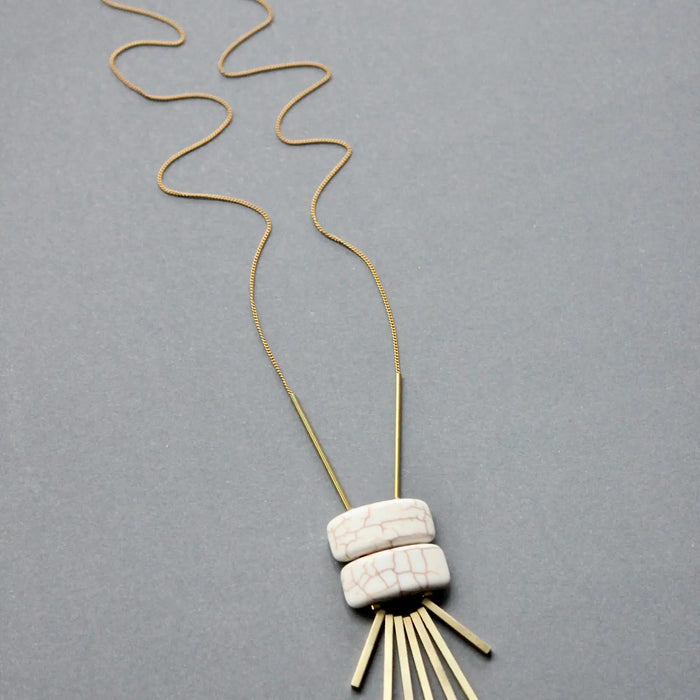 The DAVID AUBREY - WHITE GEOMETRIC PENDANT NECKLACE by DAVID AUBREY showcases two white magnesite beads above six gold-plated brass bars arranged like rays, all set against a simple gray background.