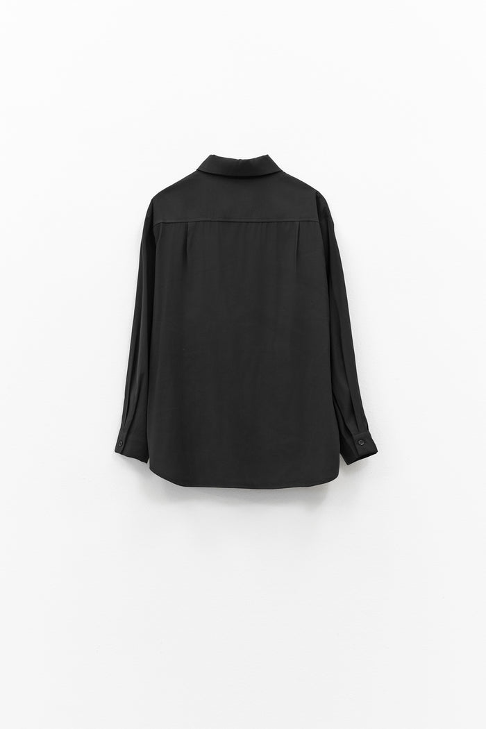 The ELEH - THE ESSENTIAL BLACK SHIRT, featuring long sleeves, a pointed collar, and chest pocket, hangs against a white background—ideal for those who value minimalist luxury.