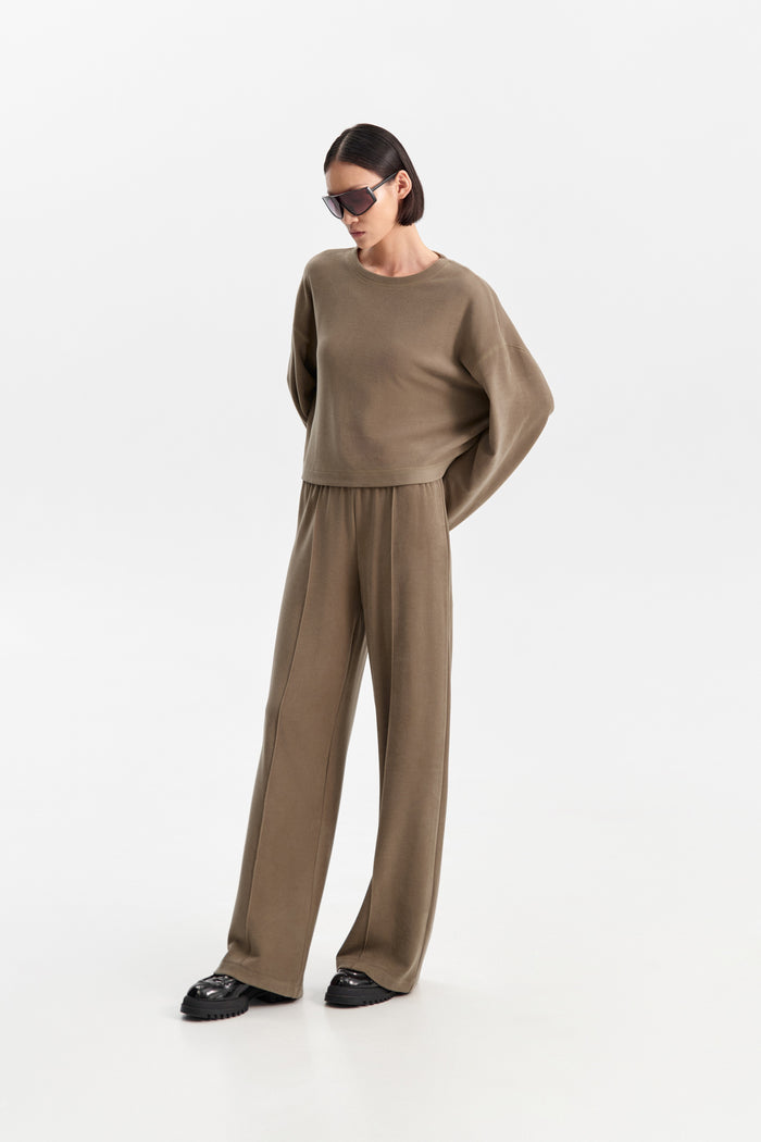 The ELEH - THE SCALA PULL ON SWEATPANT TROUSER by ELEH is a pair of loose-fitting, light brown, velvety pull-on trousers with an elastic waistband, shown against a plain white background.