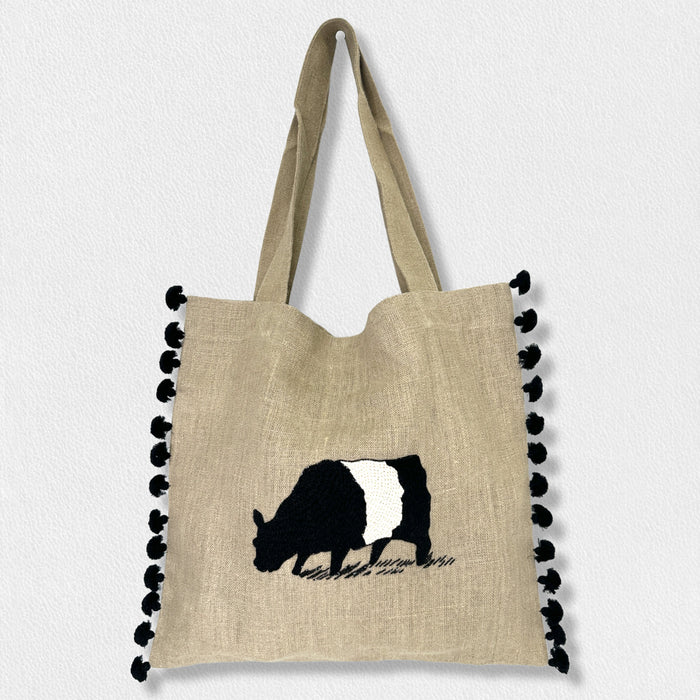 The FEARRINGTON LIFESTYLE COLLECTION - SIGNATURE BELTIE TOTE by CITRUS FASHIONS is a beige hand-stitched bag with black pom-pom trim and an embroidered cow, featuring two handles—perfect for everyday errands.