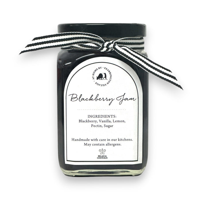 The FEARRINGTON SIGNATURE COLLECTION - FEARRINGTON HOUSEMADE BLACKBERRY JAM features a stylish black and white striped ribbon tied around the lid. The label reveals ingredients including blackberry, vanilla, lemon, pectin, and sugar. This handcrafted jam is made with meticulous care and is an ideal hostess gift. Please be aware that it may contain allergens.