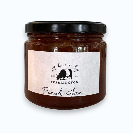 The FEARRINGTON SIGNATURE COLLECTION - FEARRINGTON HOUSEMADE PEACH JAM comes in a glass jar featuring a black lid and is adorned with a black and white striped ribbon. The white label reads "Peach Jam" and lists the ingredients as peach, vanilla, lemon, pectin, and sugar. Perfect as a hostess gift, it also notes that it is handmade by FEARRINGTON HOUSE and may contain allergens.