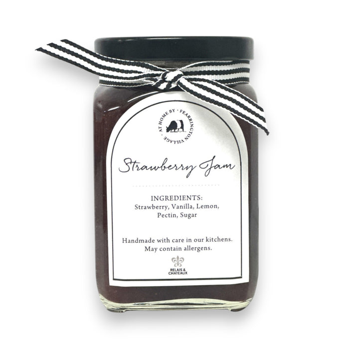 A glass jar of raspberry jam is displayed with a black and white striped ribbon adorning the lid. The white label reads "Fearrington Housemade Raspberry Jam" with a list of ingredients: raspberry, vanilla, lemon, pectin, sugar. The text also indicates it is handmade and may contain allergens. Perfect as a hostess gift!