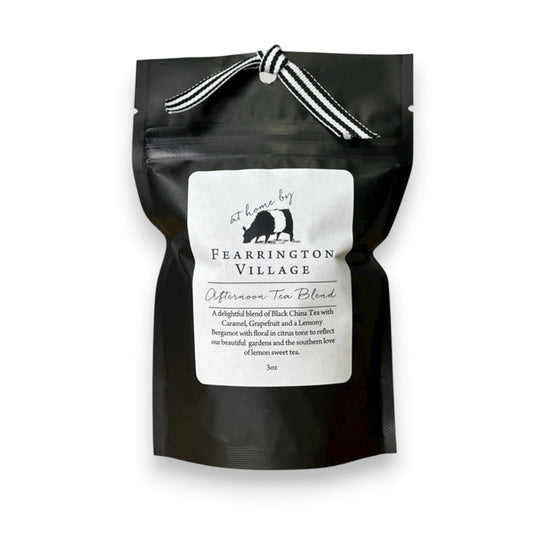 A black foil tea bag labeled "FEARRINGTON SIGNATURE COLLECTION - FEARRINGTON SIGNATURE AFTERNOON TEA BLEND" from Fearrington House, featuring a small white label with an image of a bear holding a bird. The bag contains loose leaf tea and is tied at the top with a black and white striped ribbon. It rests on a light gray surface.