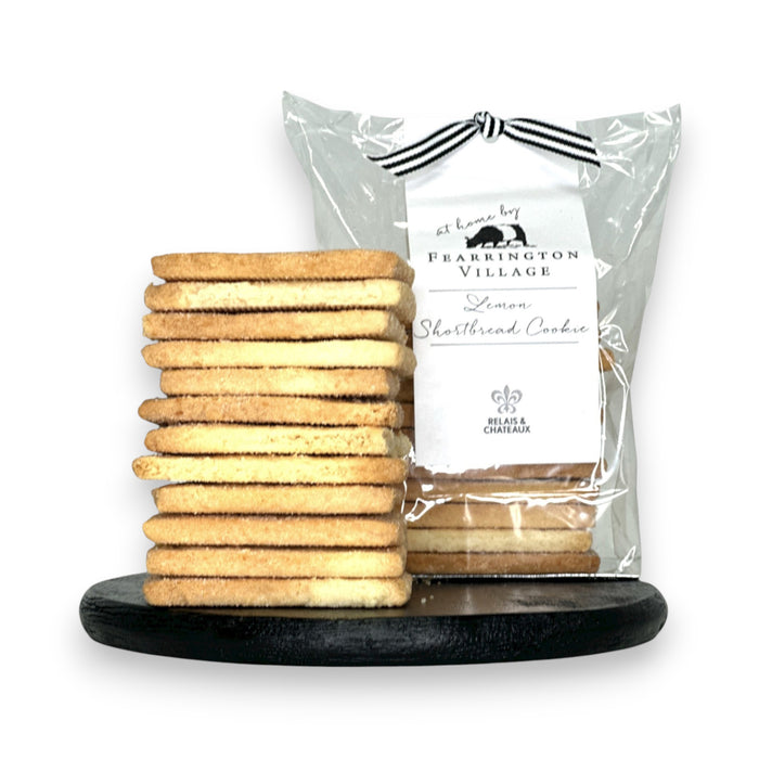 A stack of buttery, crisp FEARRINGTON SIGNATURE COLLECTION - HOUSEMADE LEMON SHORTBREAD COOKIES stands on a black round surface. Behind the stack, a clear plastic bag containing more cookies is tied with a black and white ribbon and features a label reading "FEARRINGTON HOUSE Lemon Shortbread Cookie" with decorative details from The Fearrington House Restaurant.