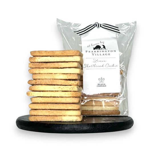 A stack of buttery, crisp FEARRINGTON SIGNATURE COLLECTION - HOUSEMADE LEMON SHORTBREAD COOKIES stands on a black round surface. Behind the stack, a clear plastic bag containing more cookies is tied with a black and white ribbon and features a label reading "FEARRINGTON HOUSE Lemon Shortbread Cookie" with decorative details from The Fearrington House Restaurant.