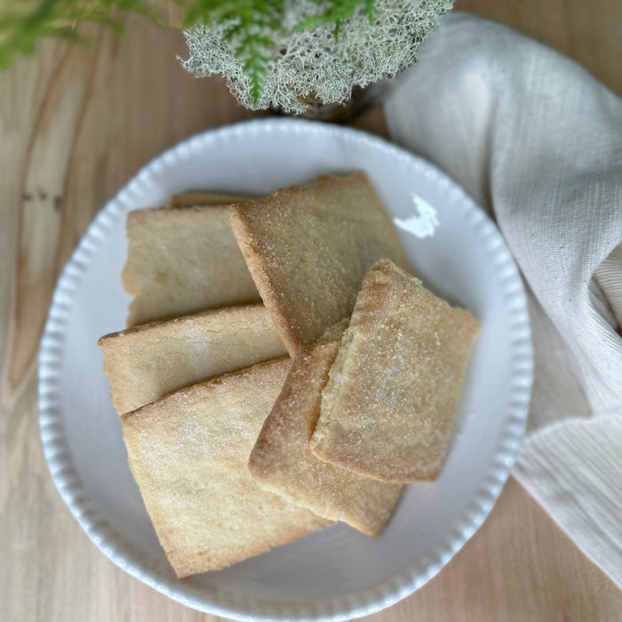 A white plate with rectangular, golden-brown FEARRINGTON HOUSE Fearrington Signature Collection - Housemade Lemon Shortbread Cookies sits on a wooden surface beside a beige cloth and a small green plant.