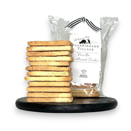 A stack of buttery and crisp vanilla shortbread cookies is displayed on a black serving board in front of a clear package labeled "Fearrington House Signature Collection - Housemade Vanilla Shortbread Cookies" with a black and white striped ribbon tie at the top. These delectable treats could easily be found at The Fearrington House Restaurant.