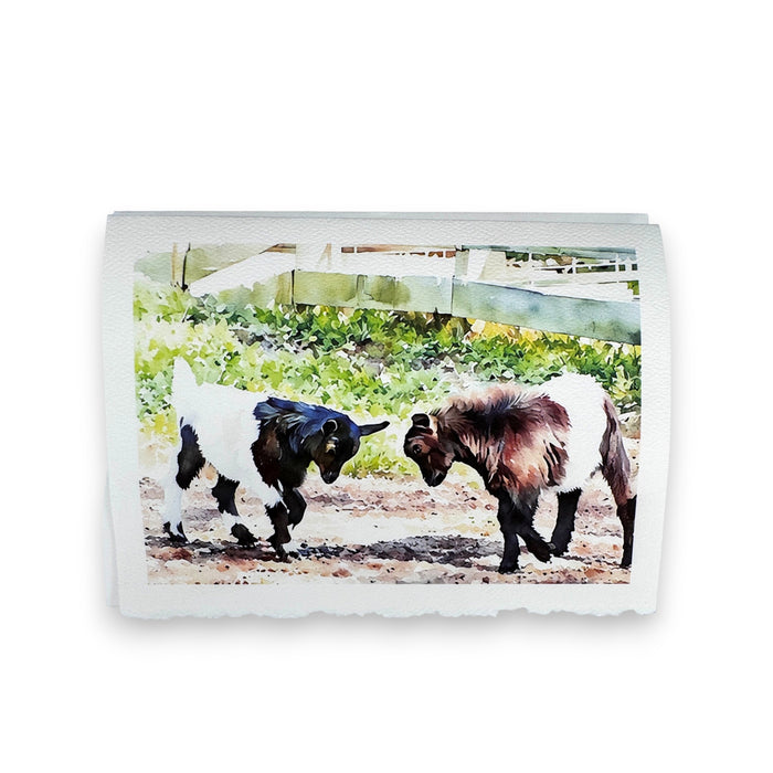 Two goats with white and brown fur playfully butting heads near a white fence on a sunny day capture the charm of Fearrington Village, featured in the PICTURE SALON FEARRINGTON SIGNATURE COLLECTION - "KNOCKING HEADS" watercolor greeting card.
