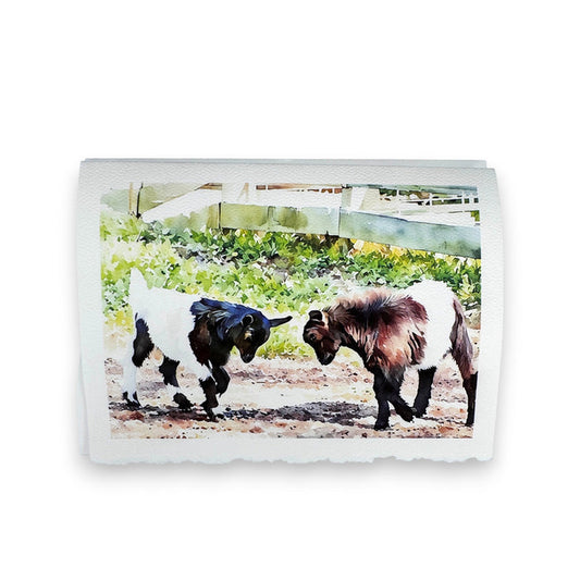 Two goats with white and brown fur playfully butting heads near a white fence on a sunny day capture the charm of Fearrington Village, featured in the PICTURE SALON FEARRINGTON SIGNATURE COLLECTION - "KNOCKING HEADS" watercolor greeting card.