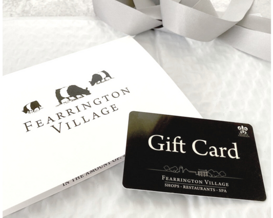 A FEARRINGTON VILLAGE GIFT CARD sits on a white envelope with the brand’s three-cow logo and a soft gray ribbon in the background. Use it at FEARRINGTON VILLAGE favorites like the Spa or Fearrington House Inn.