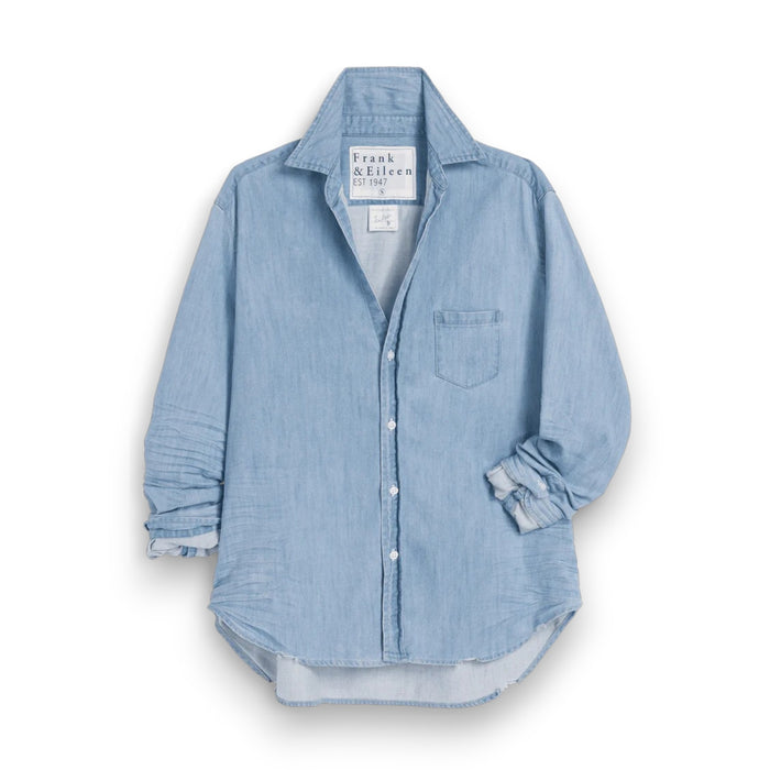 The FRANK & EILEEN Eileen Relaxed Button Up in Famous Denim Classic Wash is a light blue, long-sleeve shirt with bust-flattering buttons, a chest pocket, rolled sleeves, and a curved hem, shown against a white background.