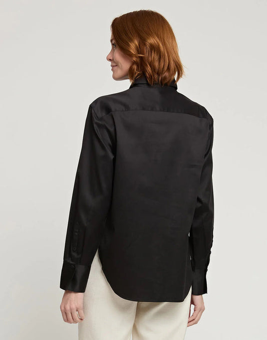 A woman with dark hair and a neutral expression wears the HINSON WU HALSEY Long Sleeve Cotton Satin Shirt with a large flower detail on the shoulder and black trousers, standing against a plain background.
