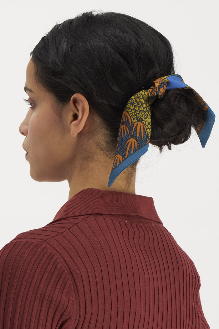 A woman with dark hair in a low bun, tied with the INOUI EDITIONS - MAGNUS SMALL SILK RIBBON by INOUI, wears a ribbed maroon top and is seen in profile against a plain white background.