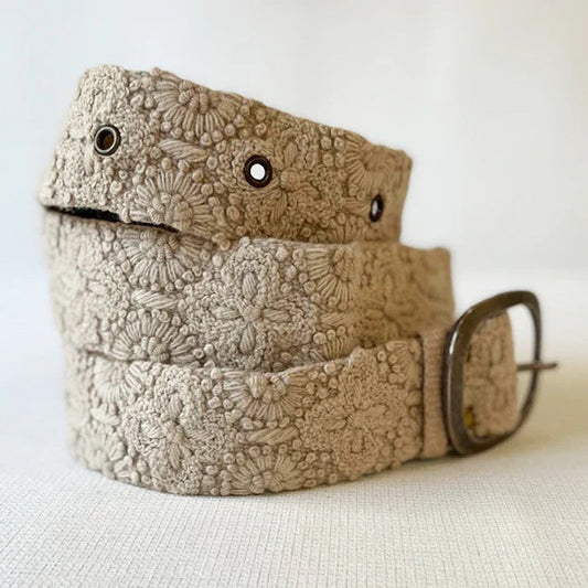The JENNY KRAUSS - DESSERT DREAMS BELT by JENNY KRAUSS is a beige, hand-embroidered Peruvian belt with intricate floral motifs, metal eyelets, and a silver buckle, artfully coiled on a light surface.