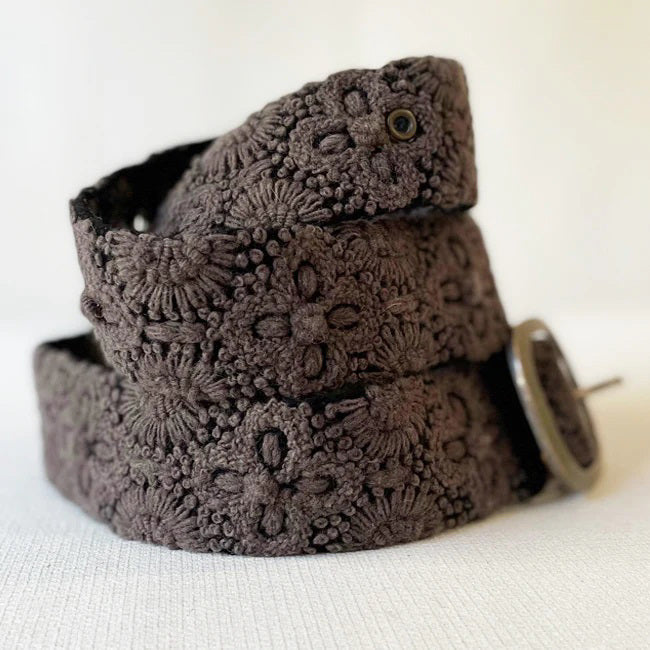 The JENNY KRAUSS - MIDNIGHT OASIS BELT features intricate raised floral embroidery, a round metal buckle, and a dark brown design, handcrafted in Peru and shown coiled on a light background.
