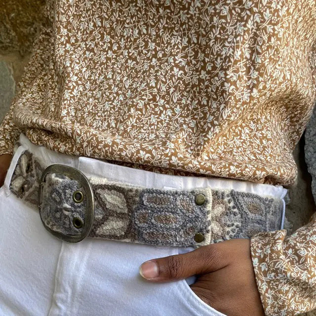 The JENNY KRAUSS Monochromatic Belt in Cream, handcrafted in Peru with floral and geometric embroidery in beige, brown, and gray, features a metal buckle and is shown coiled on a light surface.