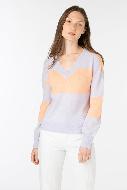 A woman with long brown hair models the KINROSS CASHMERE Gathered Sleeve Vee Neck pullover in pastel purple and peach, paired with white pants against a plain white background.