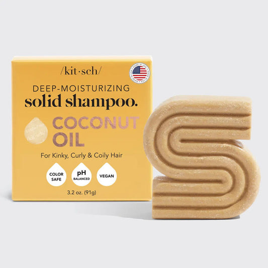 The KITSCH - Coconut Oil Deep Moisturizing Shampoo Bar, ideal for curly and kinky hair, is displayed alongside a coconut half, palm leaf, herbs, and a sample on a beige surface. The packaging highlights the inclusion of coconut oil as a key ingredient.