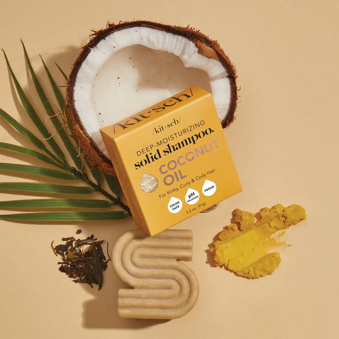 A solid shampoo bar, shaped like an "S," rests beside a yellow box labeled "KITSCH - Coconut Oil Deep Moisturizing Shampoo Bar." Ideal for curly and kinky hair, it's infused with coconut oil and boasts color-safe, pH balanced, and vegan-friendly benefits.