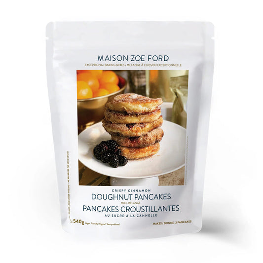 A transparent bag of MAISON ZOE FORD - CRISPY CINNAMON DOUGHUT PANCAKE MIX. The front of the bag showcases an image of several round, golden-brown pancakes stacked on a white plate with blackberries, set against a background featuring orange juice and a window.