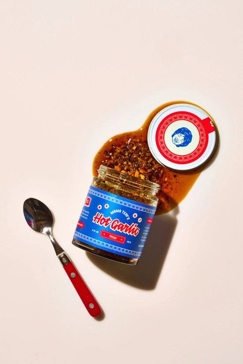 A glass jar of MAMA TEAV’S - MILD HOT GARLIC CHILI CRISPS, featuring a blue and red label, rests on a light gray background. Inside the jar is a dark, chunky condiment from MAMA TEAV’S that’s ideal for anyone looking for a vegan and gluten-free hot garlic chili crisp.
