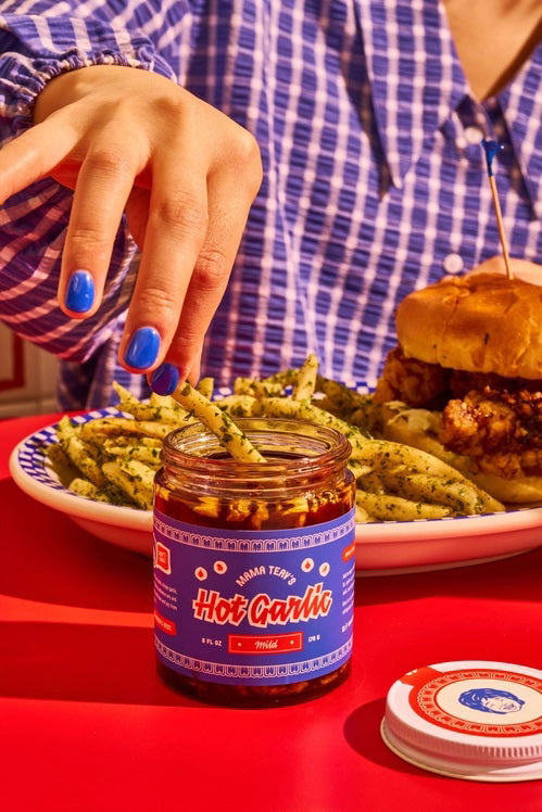 A glass jar of MAMA TEAV’S - MILD HOT GARLIC CHILI CRISPS, featuring a blue and red label, rests on a light gray background. Inside the jar is a dark, chunky condiment from MAMA TEAV’S that’s ideal for anyone looking for a vegan and gluten-free hot garlic chili crisp.