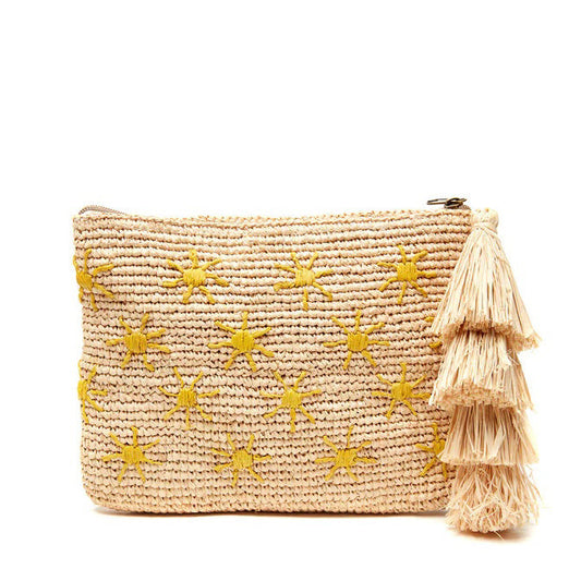 The MAR Y SOL - SOLEIL CLUTCH by MAR Y SOL, featuring yellow sun embroidery and an oversized tassel, rests on a light cloth with three lemons and brown sunglasses. Sunlight casts playful shadows across this stylish display.