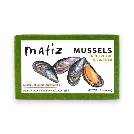 A green box of MATIZ ESPANA - MATIZ Mussels in Extra Virgin Olive Oil highlights its omega-3 content and origin from Galicia, Spain, with illustrations of Spanish mussels. Net weight: 111g.