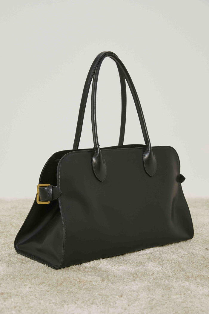 A person in loose cream pants and a matching sweater stands on carpet, holding the MEIMEIJ - BLACK LEATHER HANDBAG by its handles, adding a structured silhouette to the relaxed MEIMEIJ ensemble.