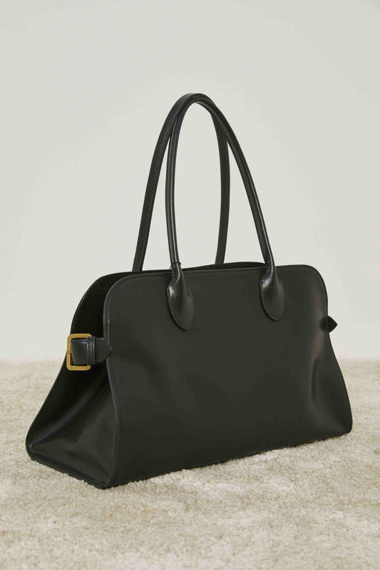 A person in loose cream pants and a matching sweater stands on carpet, holding the MEIMEIJ - BLACK LEATHER HANDBAG by its handles, adding a structured silhouette to the relaxed MEIMEIJ ensemble.