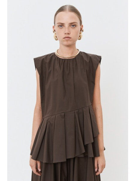 The individual is elegantly clad in the MEIMEIJ PLEATED ASYMETRICAL TOP, a sleeveless brown design with a unique peplum hem, paired with a matching long skirt. Gold hoop earrings and red flat shoes add flair against the plain white backdrop.