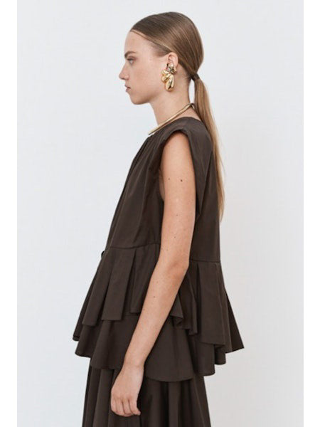 The individual is elegantly clad in the MEIMEIJ PLEATED ASYMETRICAL TOP, a sleeveless brown design with a unique peplum hem, paired with a matching long skirt. Gold hoop earrings and red flat shoes add flair against the plain white backdrop.