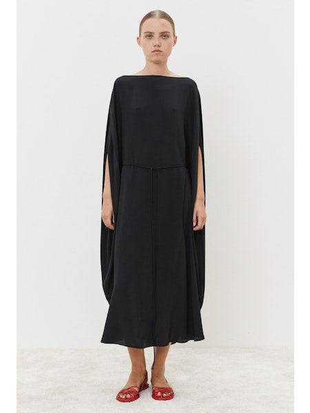 Against a white backdrop, the individual wears a MEIMEIJ Silk Blend Dress with dramatic cape-like sleeves. Their long hair flows down the elegant black gown's cinched waist, complemented by striking red sandals.