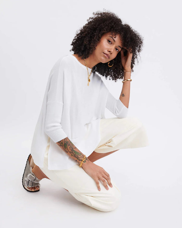 A smiling woman with curly hair wears the MERSEA - CATALINA CREWNECK SWEATER in navy blue and jeans, standing against a plain light background with her hands near her head.