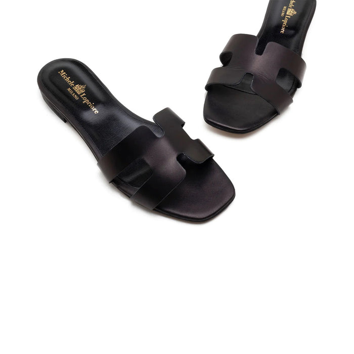 The MICHELE LOPRIORE - H SLIDE is a brown leather sandal with two wide straps, including one with a rectangular cutout, featuring a flat wooden sole. Made in Italy by MICHELE LOPRIORE, it's shown on a white background.