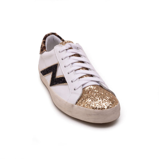 The MICHELE LOPRIORE - NINA SNEAKER features soft white leather, a black heel, glittery silver toe cap, zigzag black and beige side detailing, and a durable rubber sole, presented on a white background.