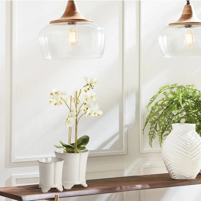 A wooden shelf holds two potted plants: a realistic NAPA HOME AND GARDEN Phalaenopsis Orchid Drop-In 20" with white orchids and another with lush green leaves. A textured white vase completes the no-maintenance décor. Above, two glass pendant lights illuminate the white paneled wall.