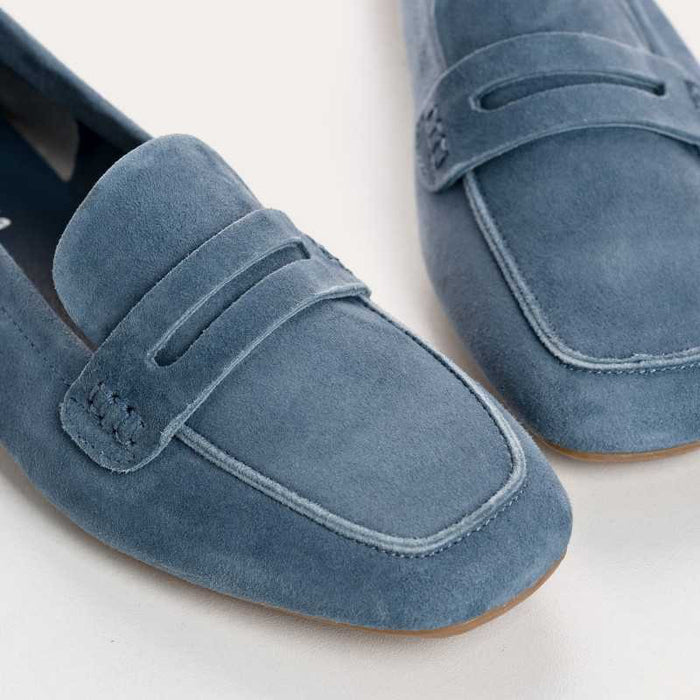 Close-up of a pair of blue suede loafers with stitching details and a penny strap, shown on a light surface.